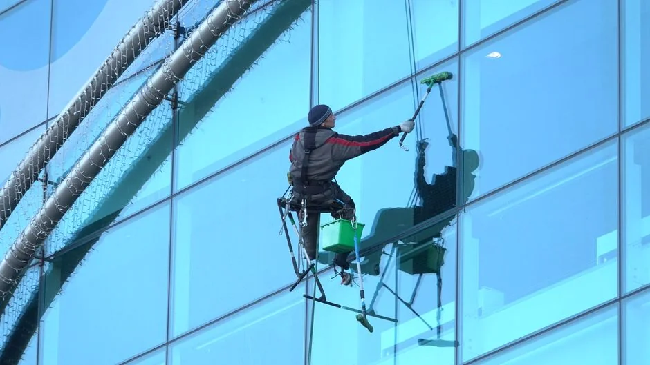 Un homme nettoie les vitres d’un gratte-ciel, harnaché, illustrant le travail en hauteur en milieu urbain.